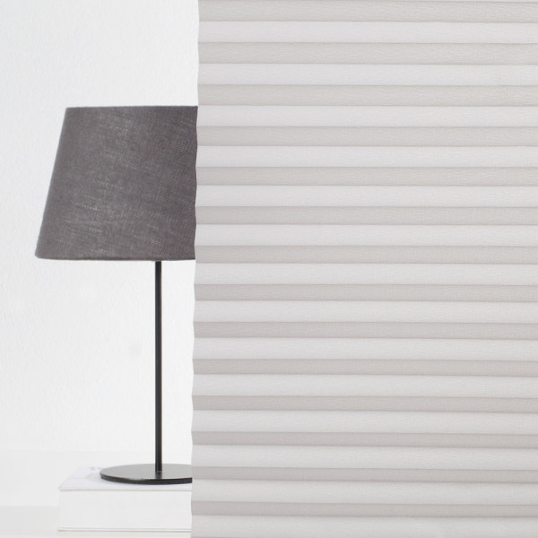 Hellgraues Plissee mit gefalteten Stoffbahnen liegt ordentlich gefaltet auf einer Oberfläche. Es ist in unterschiedlichen Maßen erhältlich und eignet sich als Sichtschutz.