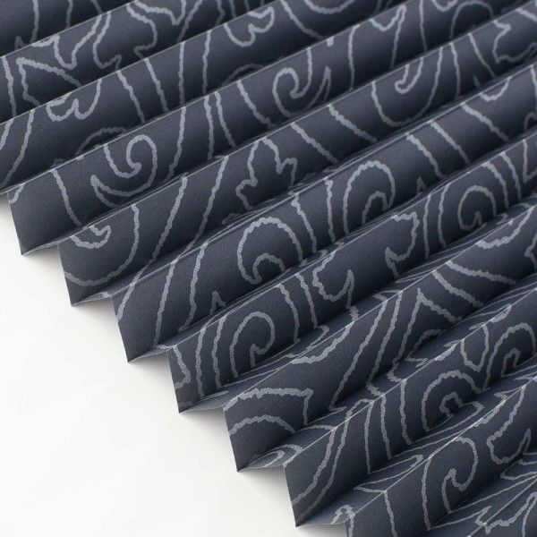 Dunkelgraues Plissee mit floralem Muster liegt gefaltet auf einem weißen Untergrund. Die Falten sind gleichmäßig angeordnet und erzeugen eine strukturierte Optik.