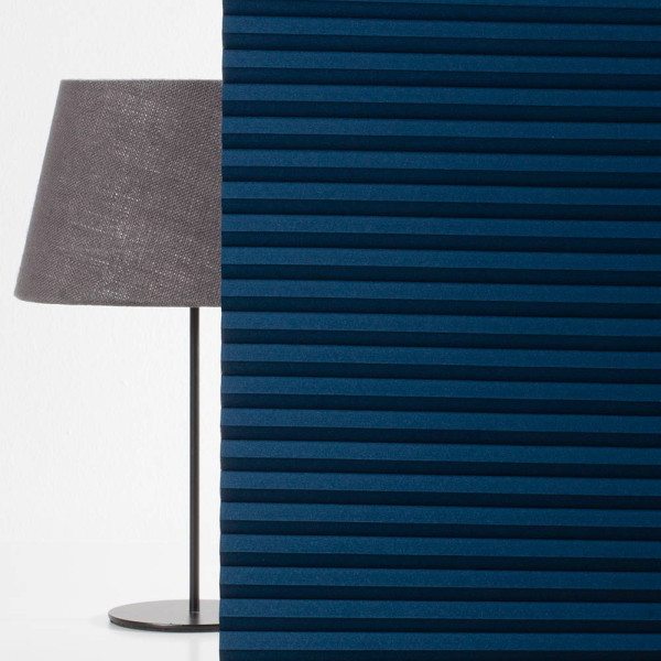 Dunkelblauer Wabenplissee-Stoff mit gerippter Struktur steht neben einer grauen Tischlampe auf einem weißen Tisch. Der Plissee ist zur Verdunkelung geeignet und individuell anpassbar.