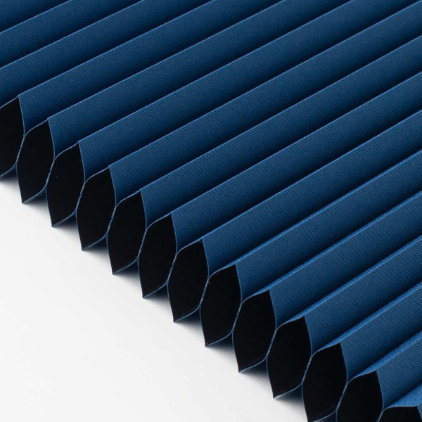 Dunkelblauer Wabenplissee-Stoff mit gerippter Struktur steht neben einer grauen Tischlampe auf einem weißen Tisch. Der Plissee ist zur Verdunkelung geeignet und individuell anpassbar.