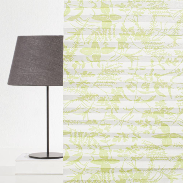 Hellgrünes, halbtransparentes Plissee mit Pflanzenmotiven in sanften Grüntönen liegt gefaltet auf einer weißen Oberfläche. Es wird zur Raumgestaltung und Lichtregulierung verwendet.