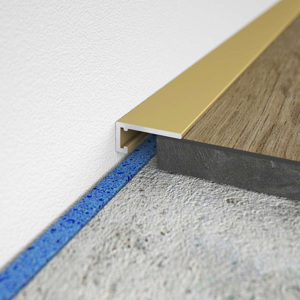 Minileiste Abschlussprofil in Gold mit 16 mm Breite und 5 mm Höhe, installiert an einer Wand mit einem Holz- und einem Steinboden. Der Bereich ist sauber und ordentlich.