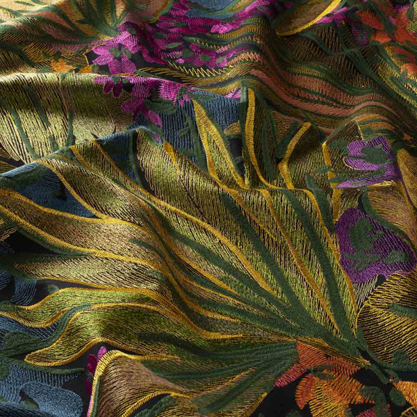 Buntes Stoffmuster mit pflanzlichem Design, featuring grüne und goldene Blätter sowie lila Blumen. Der Stoff liegt leicht gewellt und zeigt komplexe Stickereien.